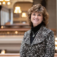 A professional portrait of a woman smiling, wearing a stylish black and white patterned blazer, set in a well-lit interior with elegant architectural features.