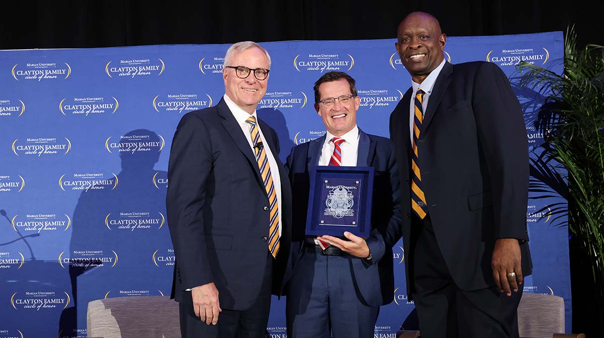 Three men in formal attire are standing together, smiling and holding an award plaque during an event. The backdrop features the Clayton's Family logo.
