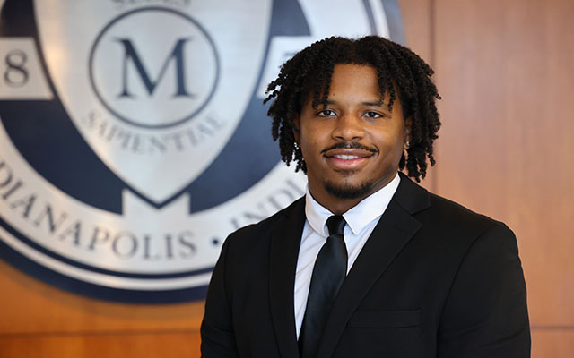 Professional portrait of Devin Sanders in a suit, smiling in front of a school logo backdrop.