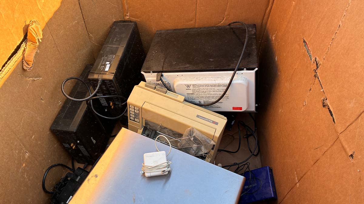 A collection of discarded electronics, including outdated computers and a microwave, inside a cardboard box.