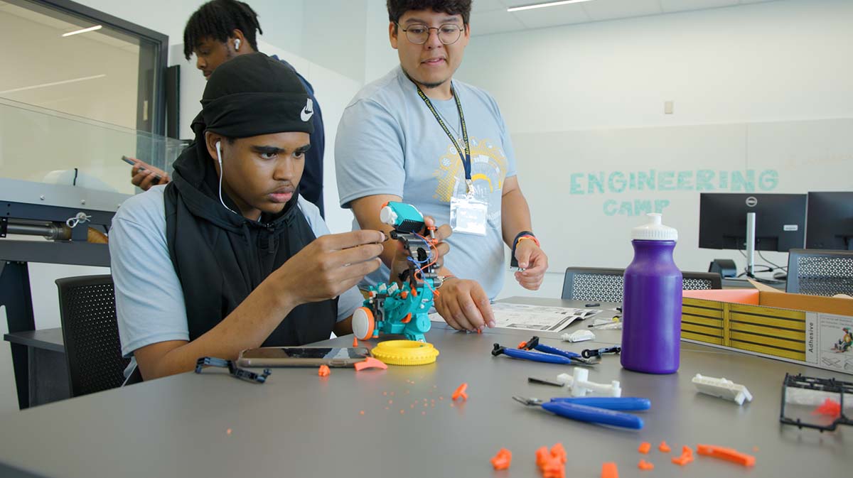 Two students engaged in hands-on activities during an engineering camp, one assembling a robotic figure while the other provides assistance.