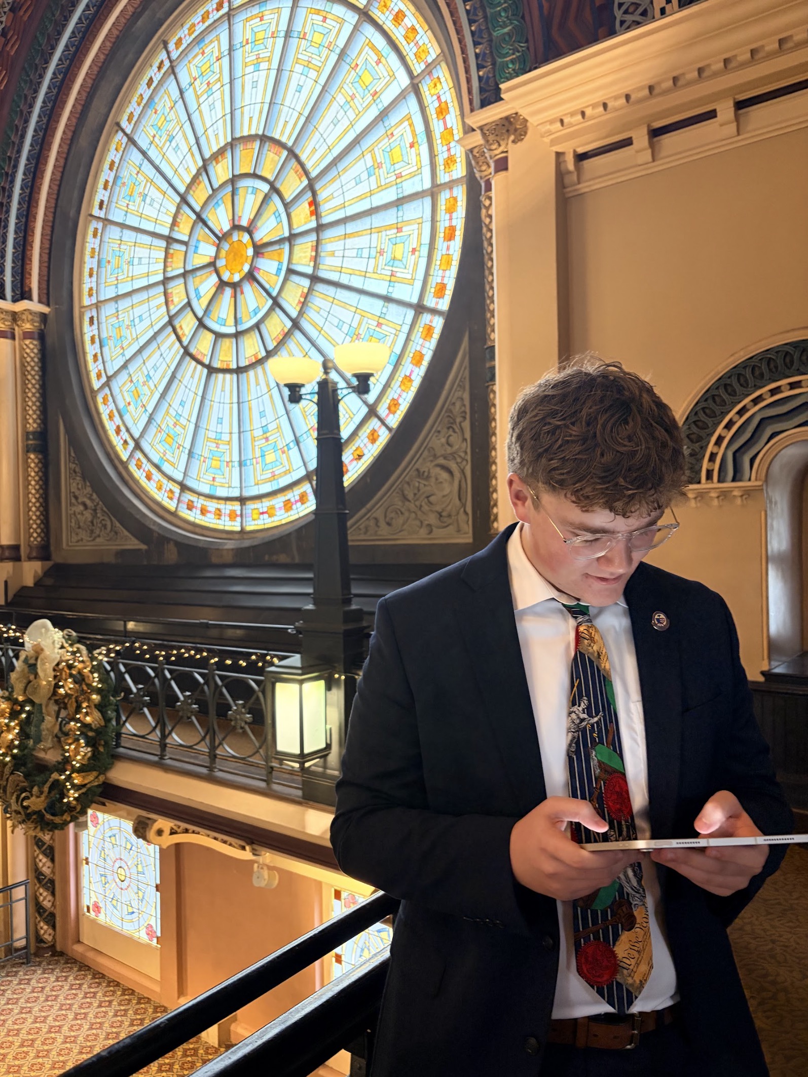 A man in a suit is using a tablet while standing near a colorful stained glass window in an ornate building.