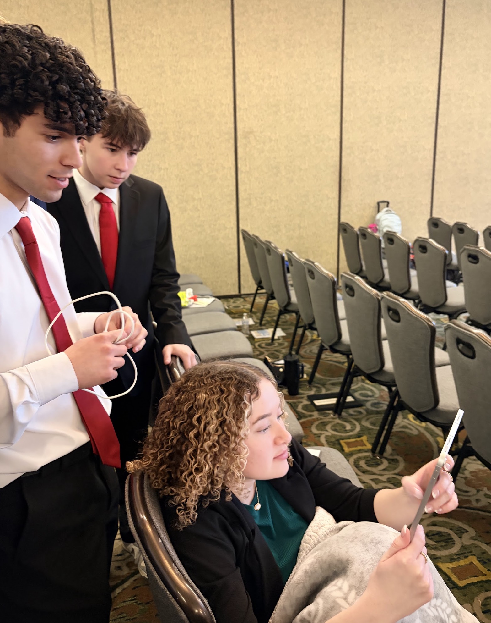 Three young individuals in formal attire engaged in a discussion, with one person seated and looking at a device, while the others stand nearby.