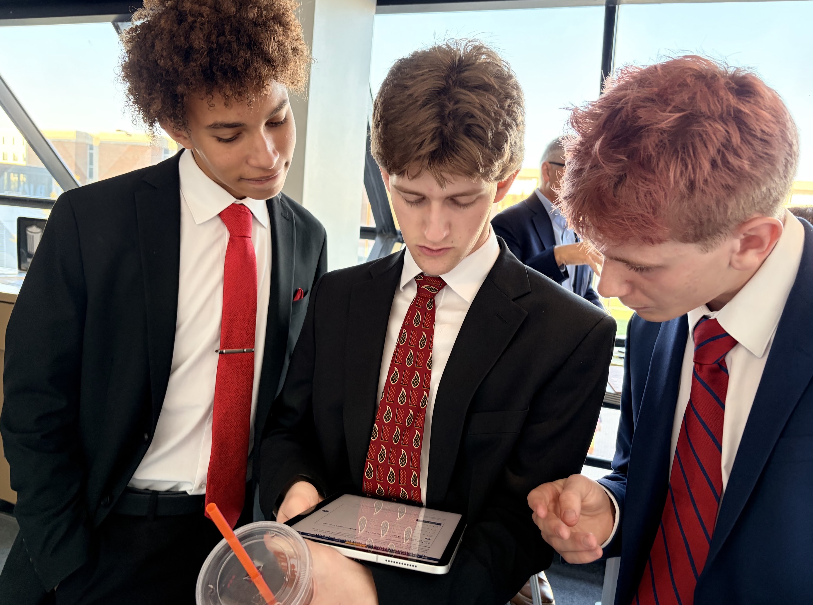 Three young men in formal attire are gathered around a tablet, engaged in discussion while looking at the screen.