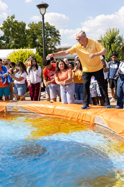 President Elsener casts something into a colorful pool while a crowd watches eagerly.