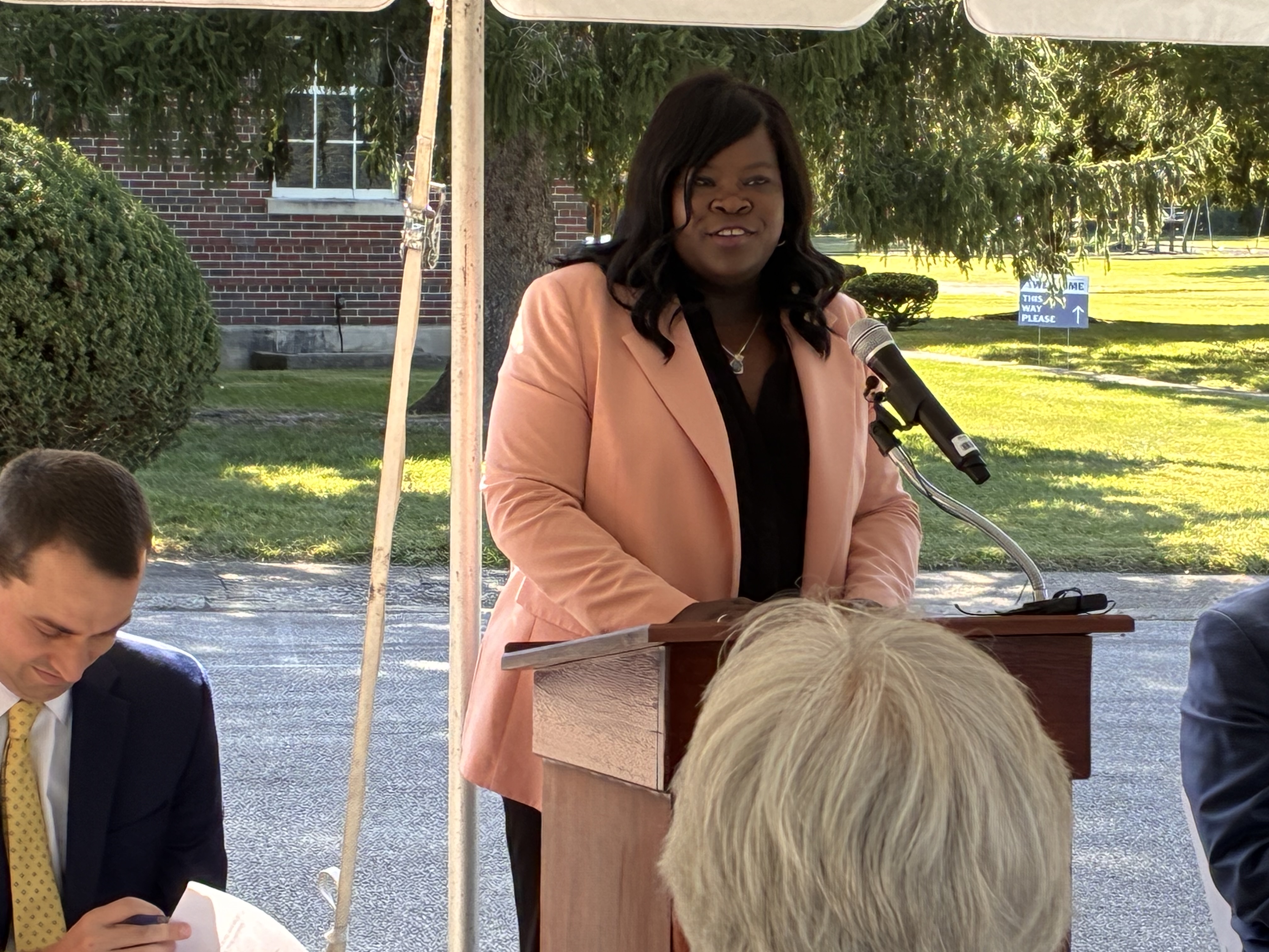A woman in an elegant pink blazer speaks at a podium during an outdoor event, with a seated audience visible in the foreground.