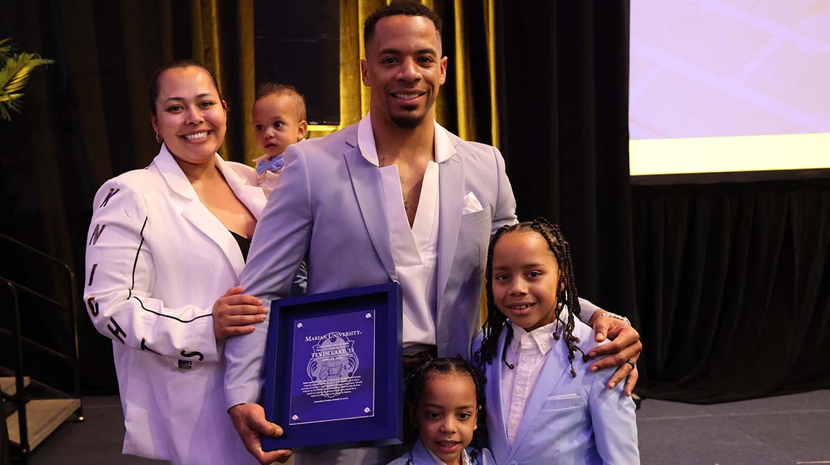 A man in a light gray suit holds an award while posing with his family, including his partner and two children, at a celebratory event.