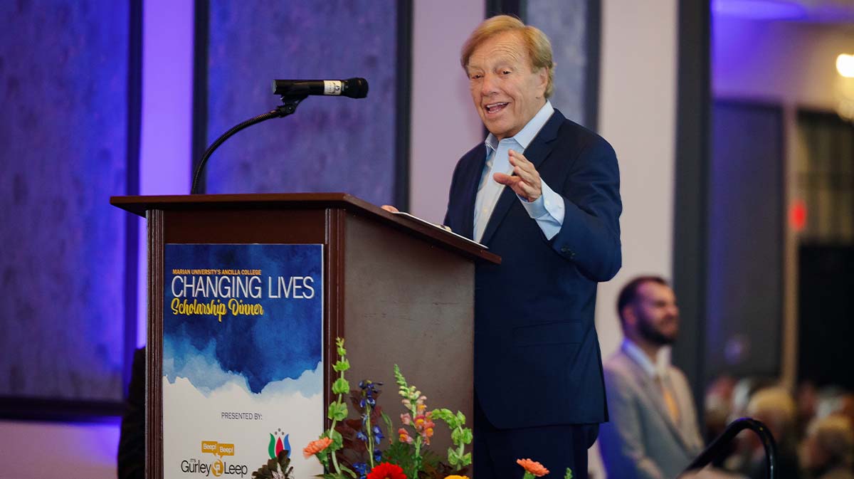 A speaker presents at the Changing Lives Scholarship Dinner, standing behind a podium adorned with event branding and floral arrangements.