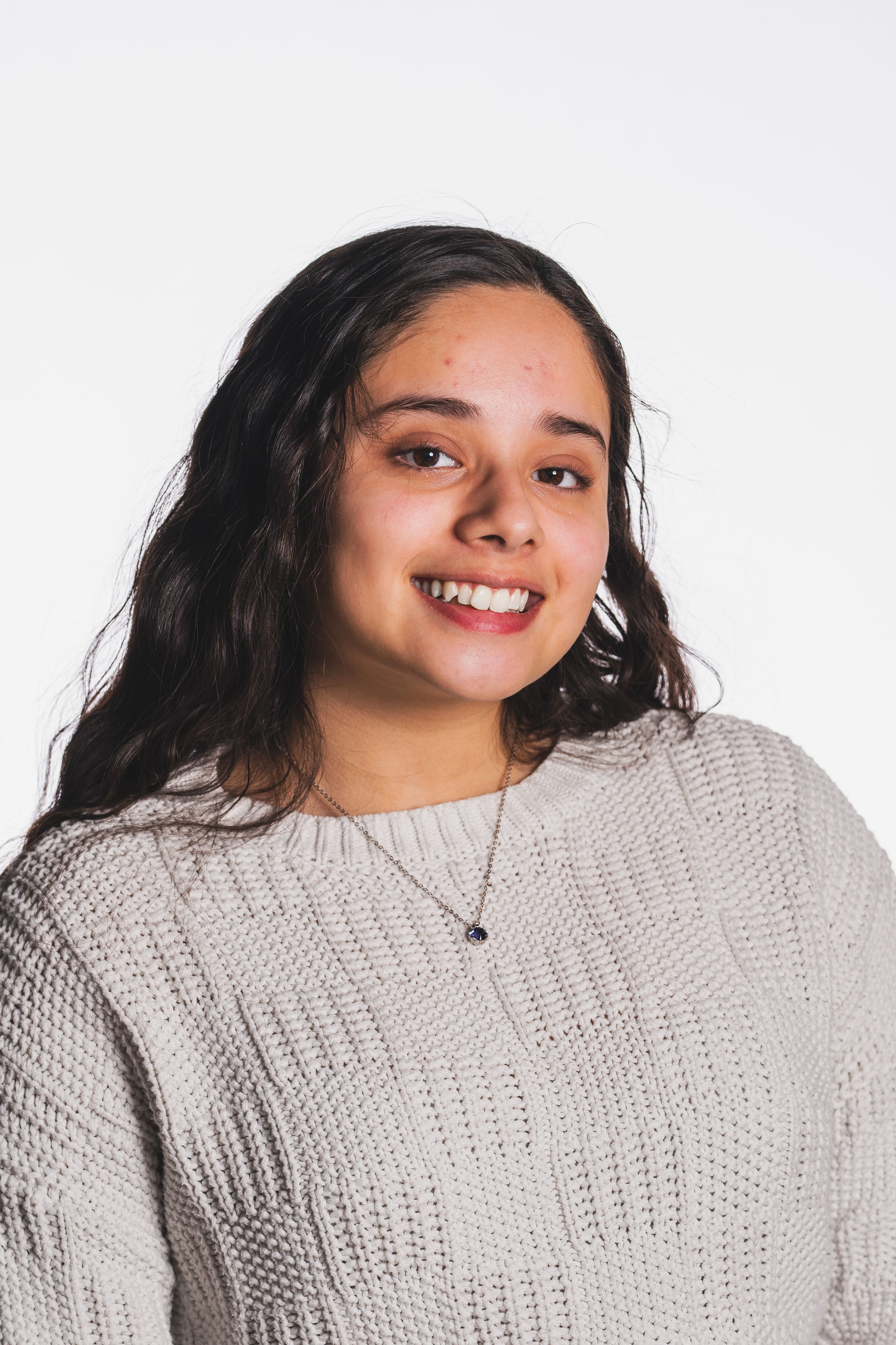 Smiling young woman with long dark hair wearing a cozy light sweater against a plain background.