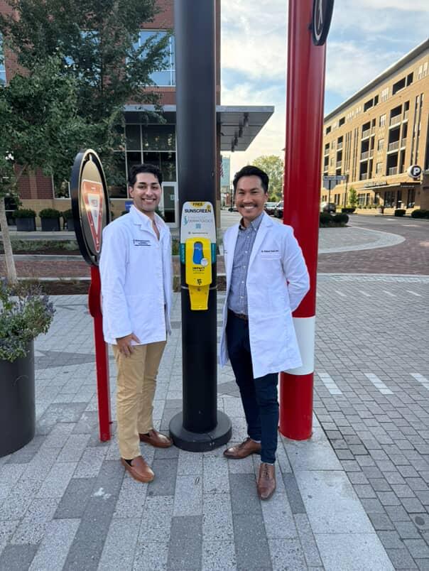 Two medical professionals in white coats stand next to a public emergency call box in an urban setting.
