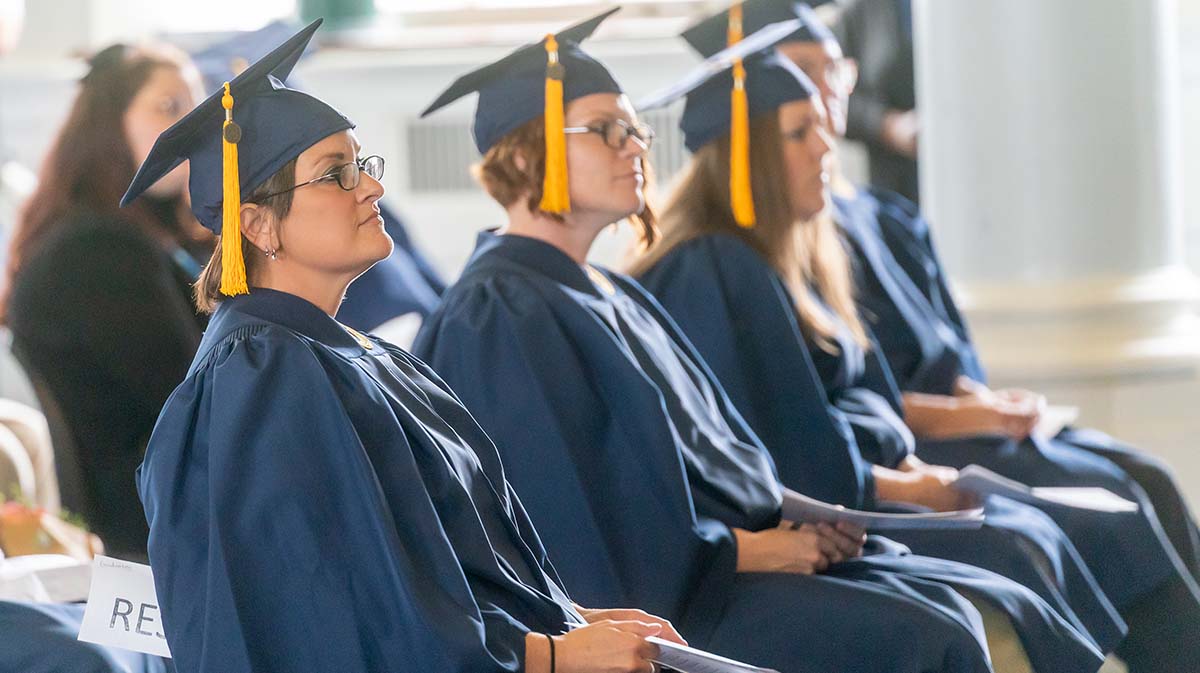 A group of graduates wearing navy blue caps and gowns attentively listening during a graduation ceremony.