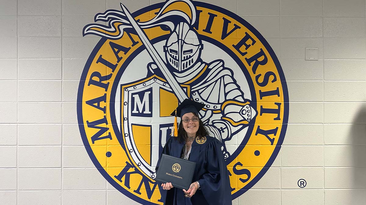 A graduate in a cap and gown proudly holds her diploma in front of the Marian University Knights logo.