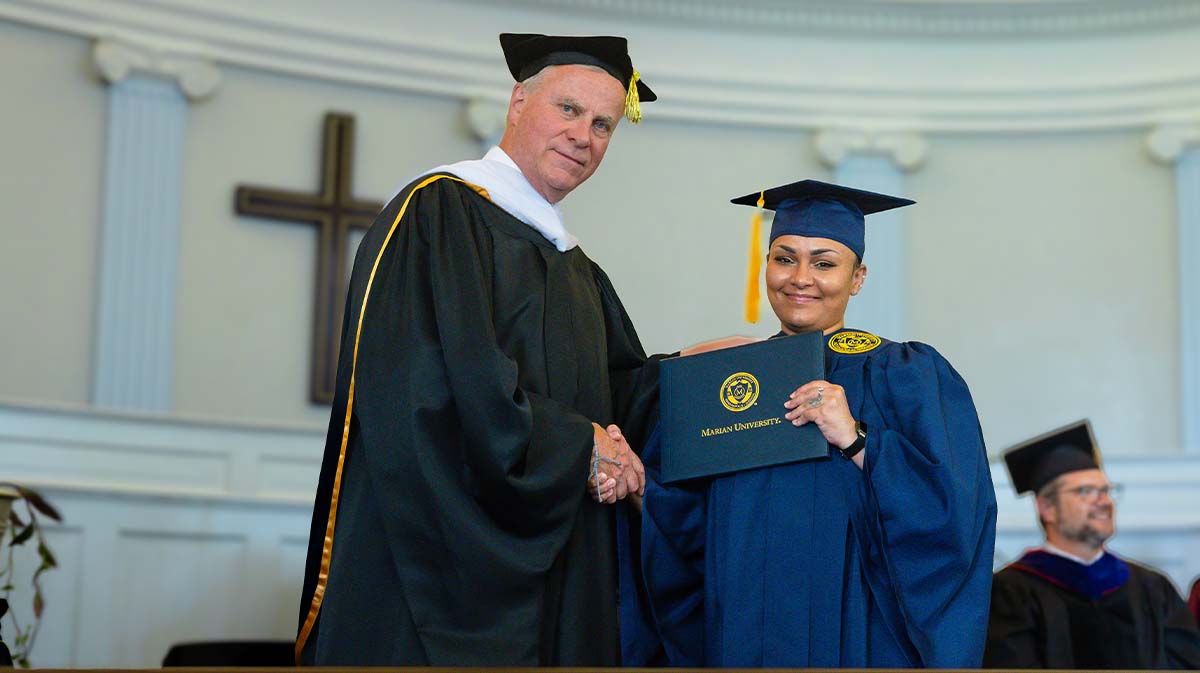 A graduate in a blue cap and gown proudly holds her diploma while shaking hands with a faculty member during a graduation ceremony.