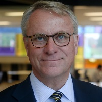 Head-and-shoulders portrait of an older man with gray hair and glasses in a suit and striped tie, with a blurred office background.