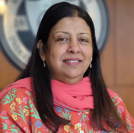 Smiling woman in a pink floral kurta and matching scarf, posed indoors with a blurred circular emblem in the background.