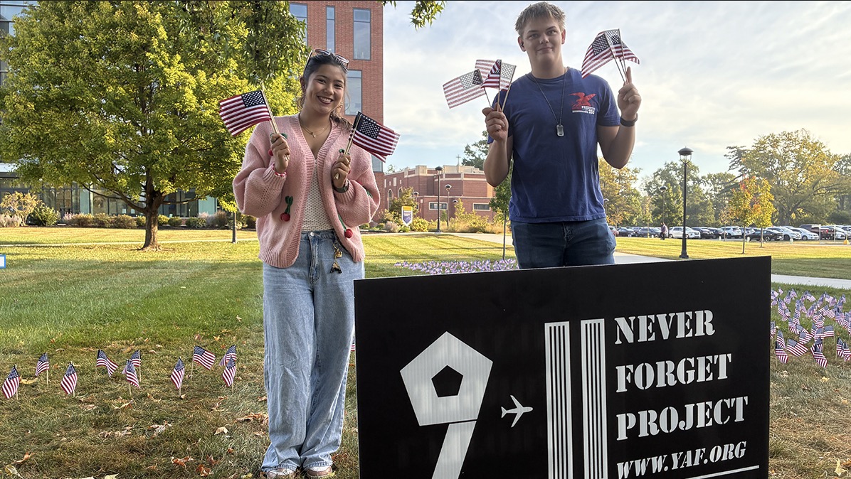 Two students hold small American flags on a campus lawn beside a large sign for the 9/11 Never Forget Project, with many flags planted in the grass behind them.