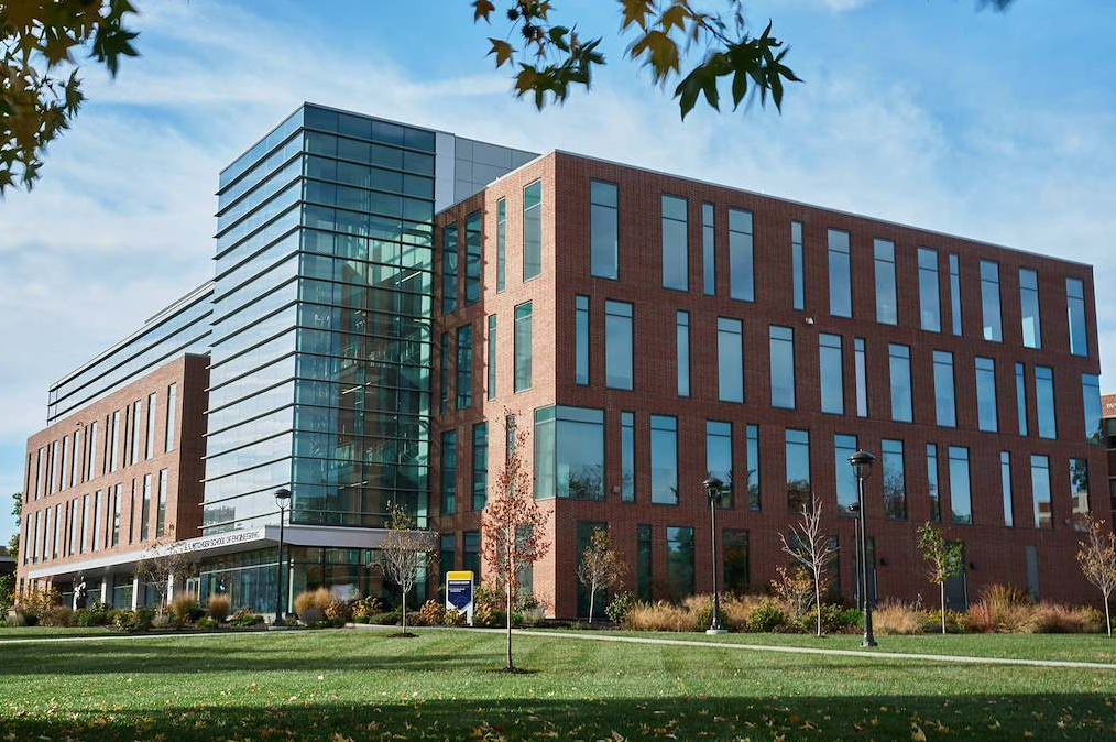 Modern multi-story academic building with a glass corner atrium and brick facade, viewed across a landscaped lawn on a clear day.