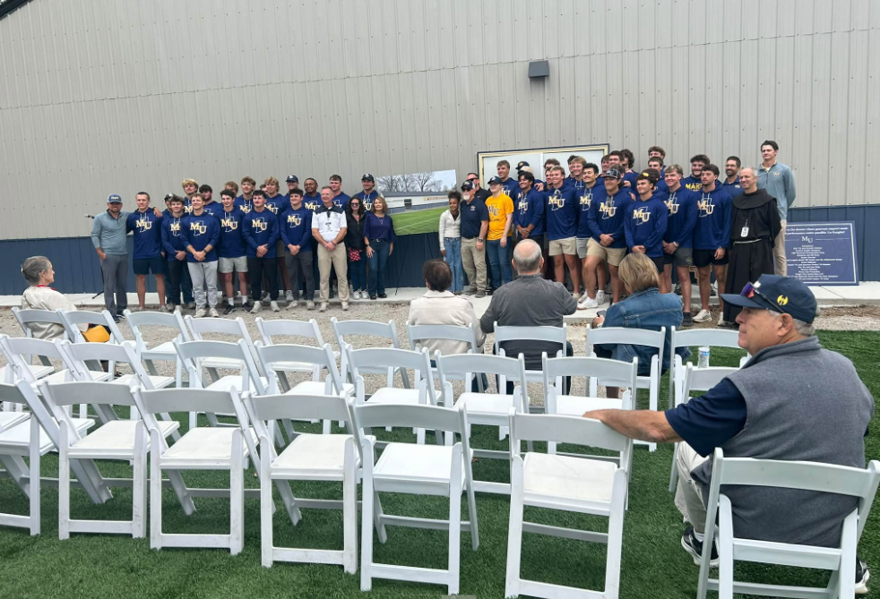 Large group of students and staff in matching MU hoodies pose for a photo in front of a building wall, with rows of white folding chairs set up in the foreground.