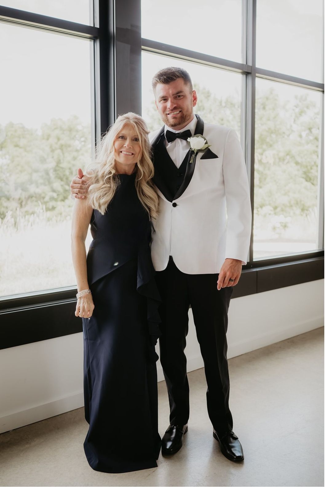 A man in a white tuxedo and black bow tie stands next to a woman in a black evening gown, both smiling in front of large windows.