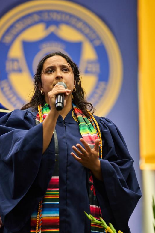 Graduation speaker in a navy cap and gown holds a microphone and gestures, with a blurred school emblem in the background.