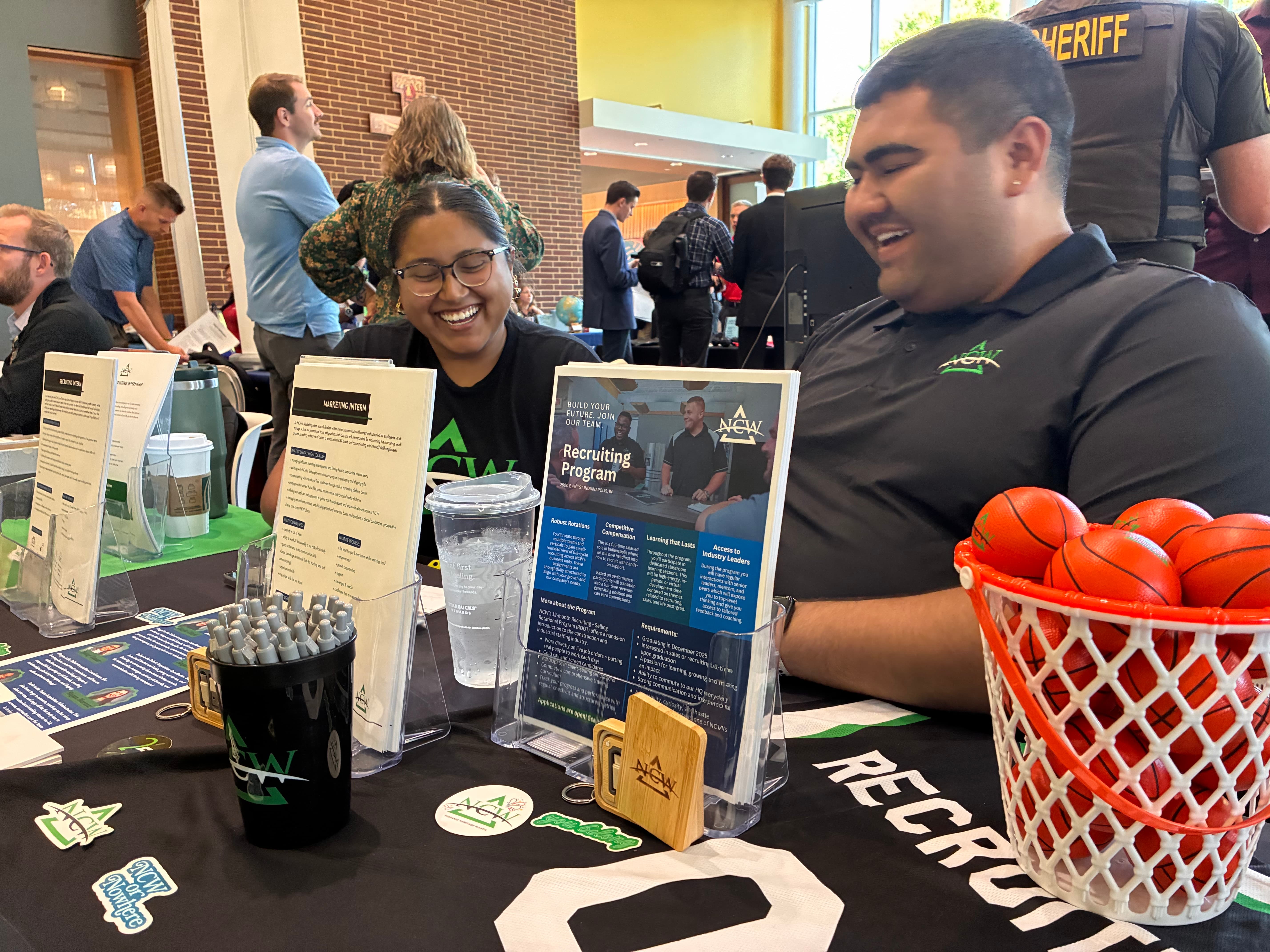 Two smiling individuals engage at a recruitment table, featuring informational materials and a basketball-themed display.