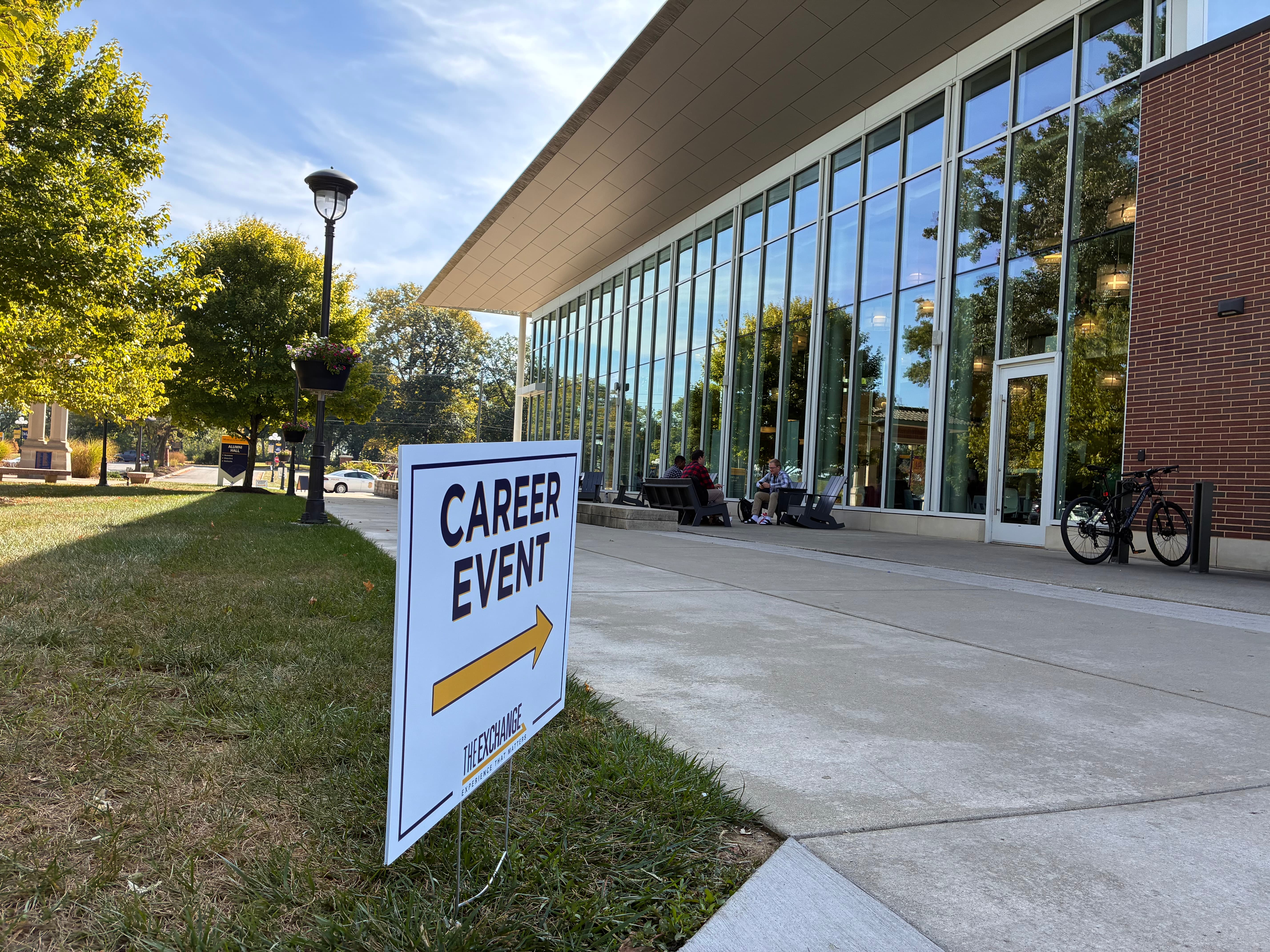 Sign for a career event pointing towards the entrance of a modern building with clear glass walls and lush green landscaping.