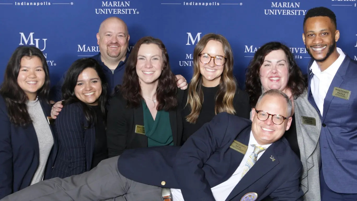 A group portrait of Marian University staff and students, smiling together in front of a backdrop featuring the university's name.