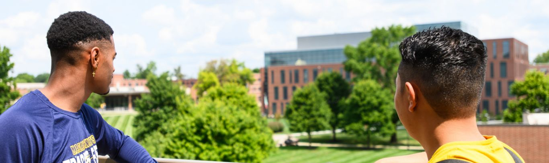 Two young men looking out over a campus landscape, featuring green trees and modern buildings in the background.