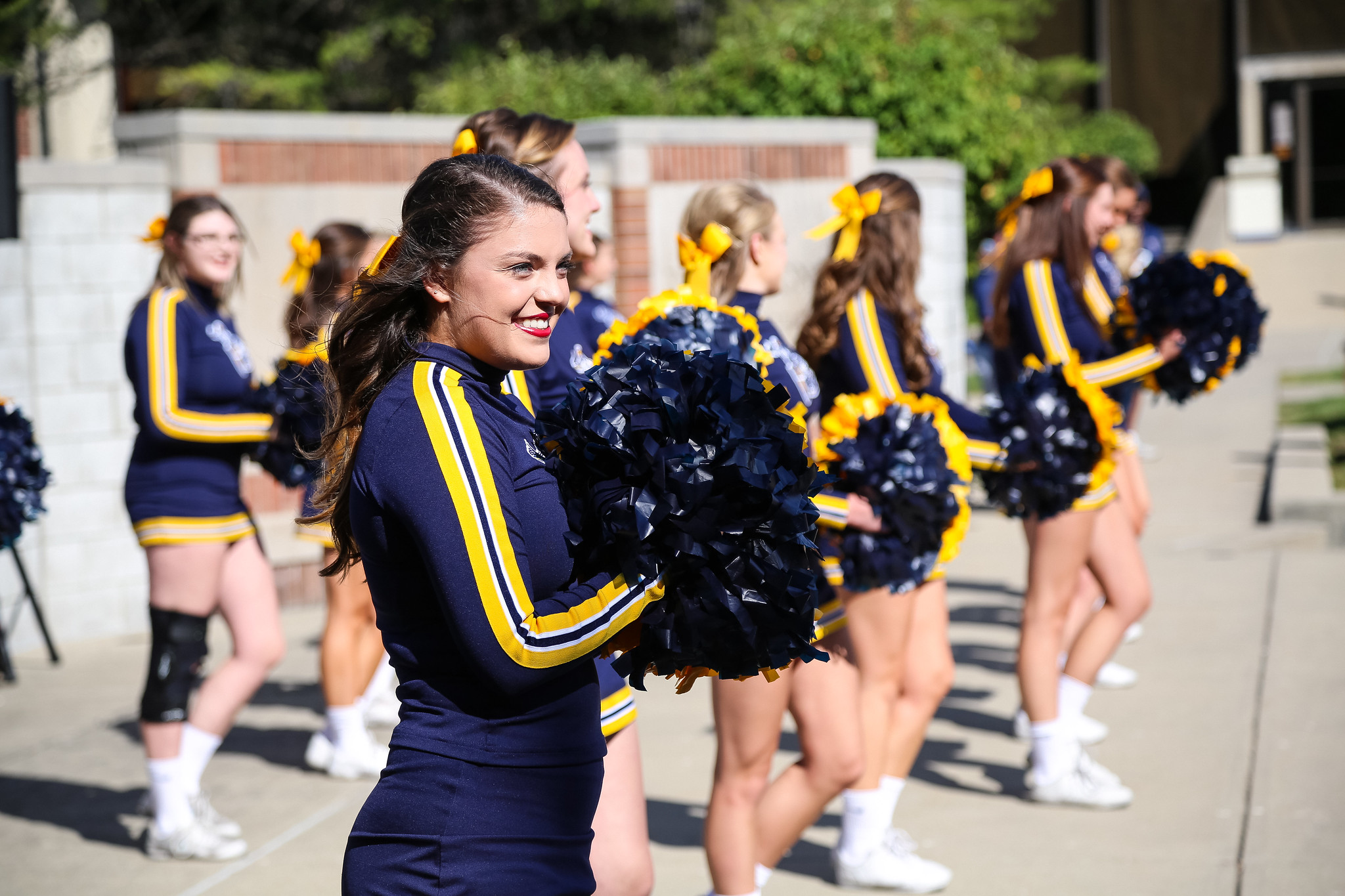 A group of cheerleaders in blue and gold uniforms are performing, with one cheerleader smiling and holding pom-poms in the foreground.