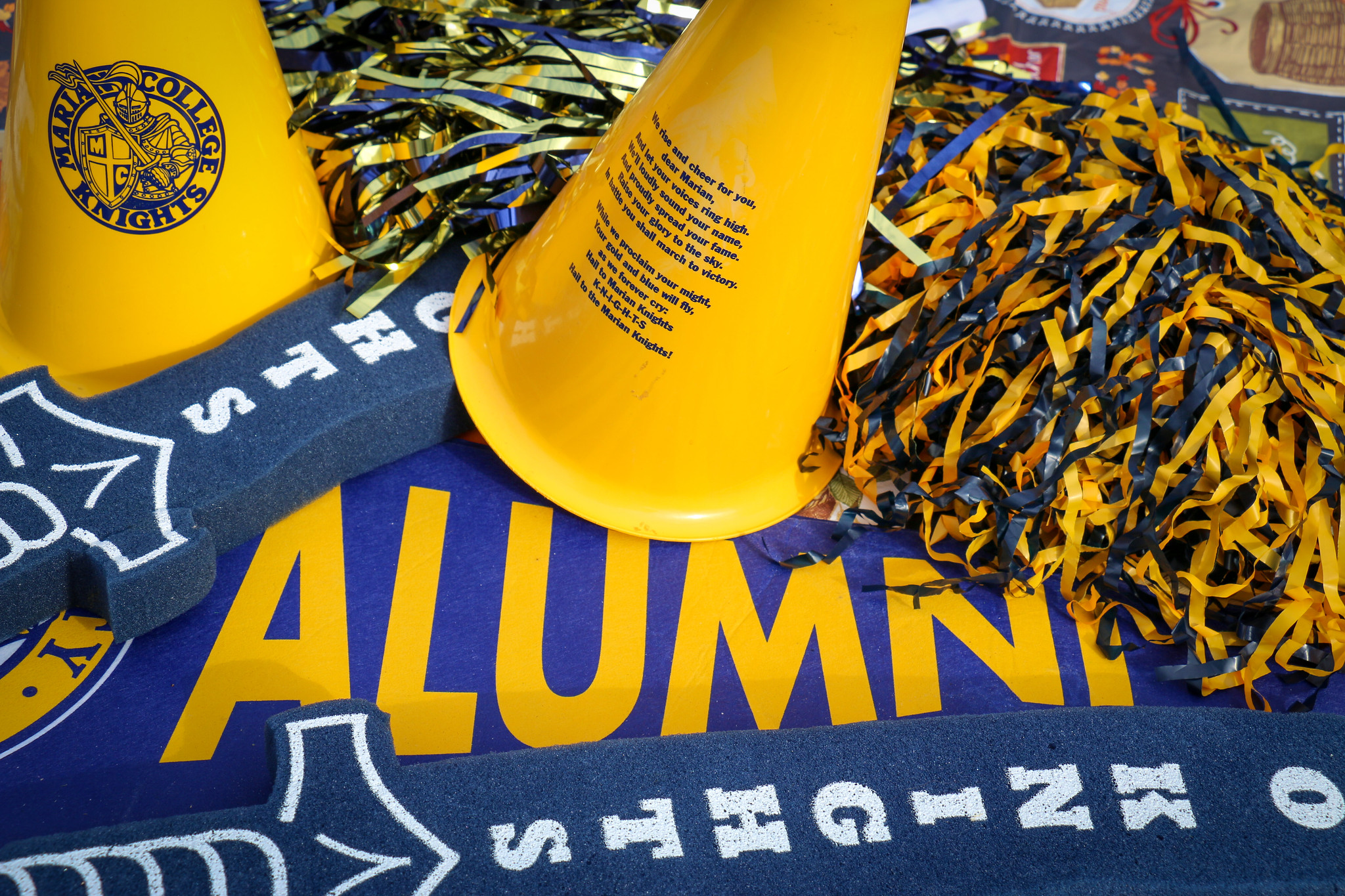 A vibrant display of alumni merchandise including yellow cheer cones, black and gold pom-poms, and a blue banner with the word "ALUMNI."