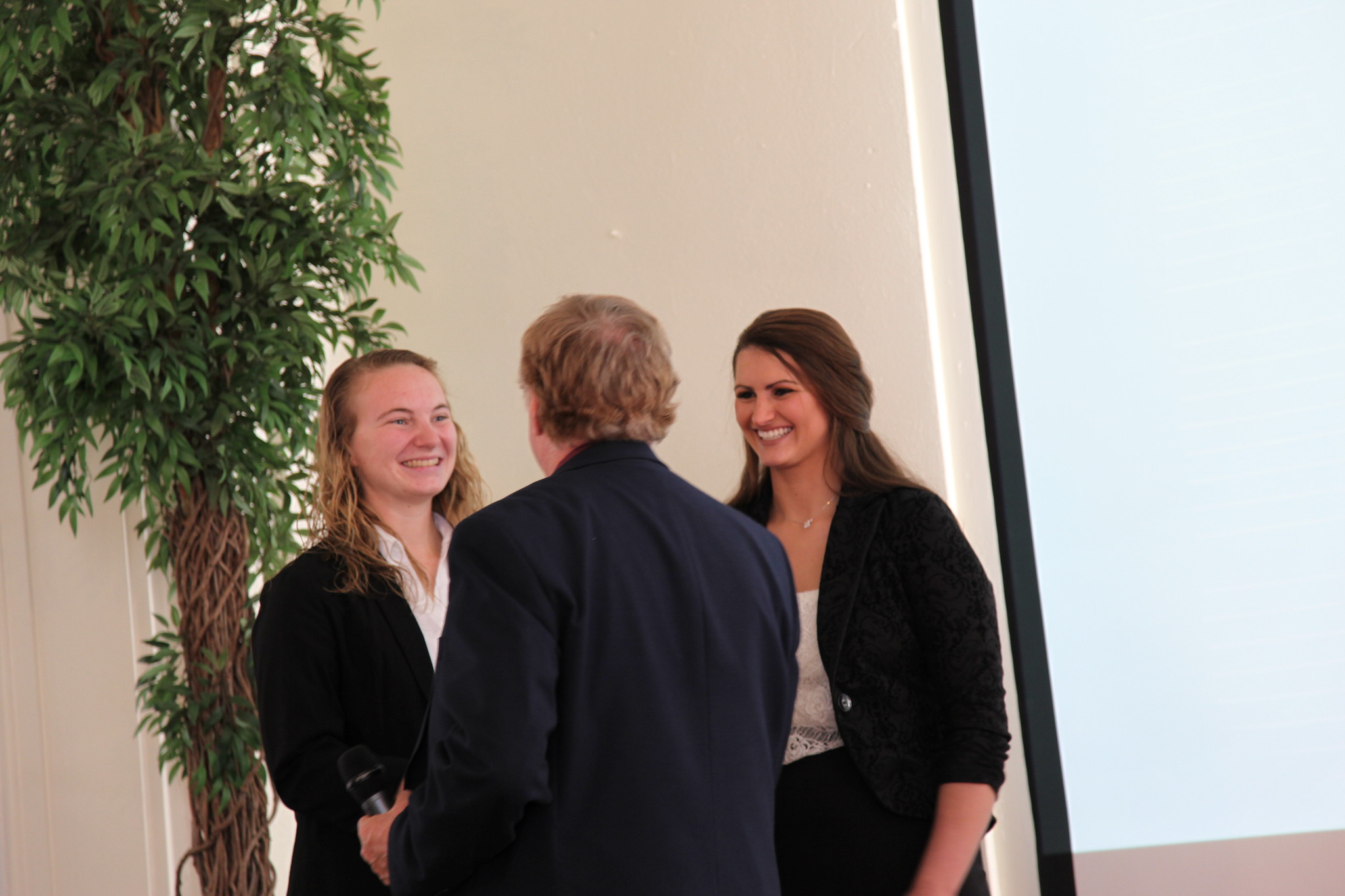 Two smiling women in professional attire are engaged in a conversation with a man, presumably during a formal event or ceremony.