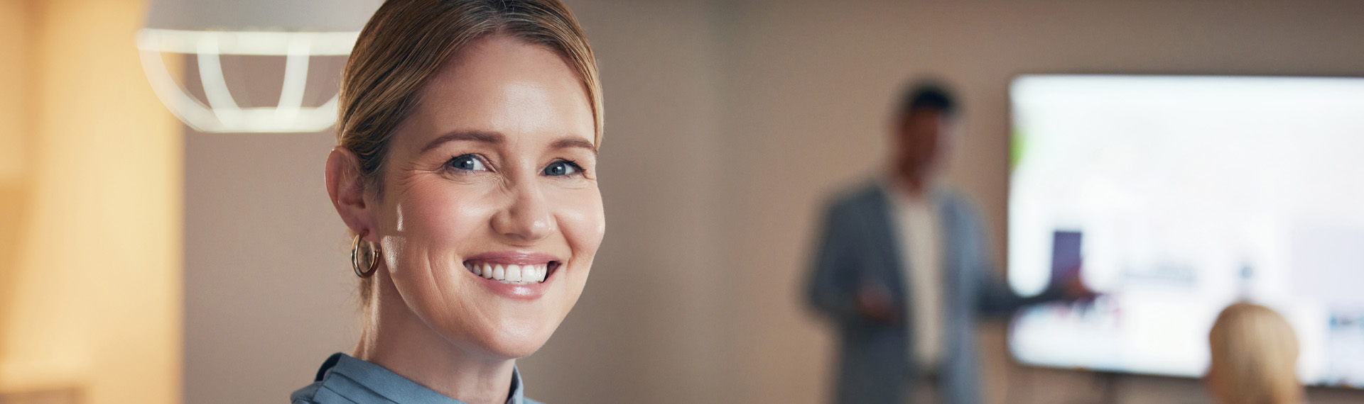 Confident smiling businesswoman in a modern office, looking directly at the camera during a professional presentation, with colleagues and a large screen in the blurred background.