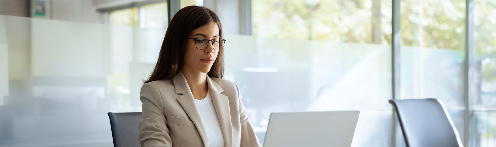 Focused young professional woman in a blazer working intently on a laptop in a modern glass-walled office.
