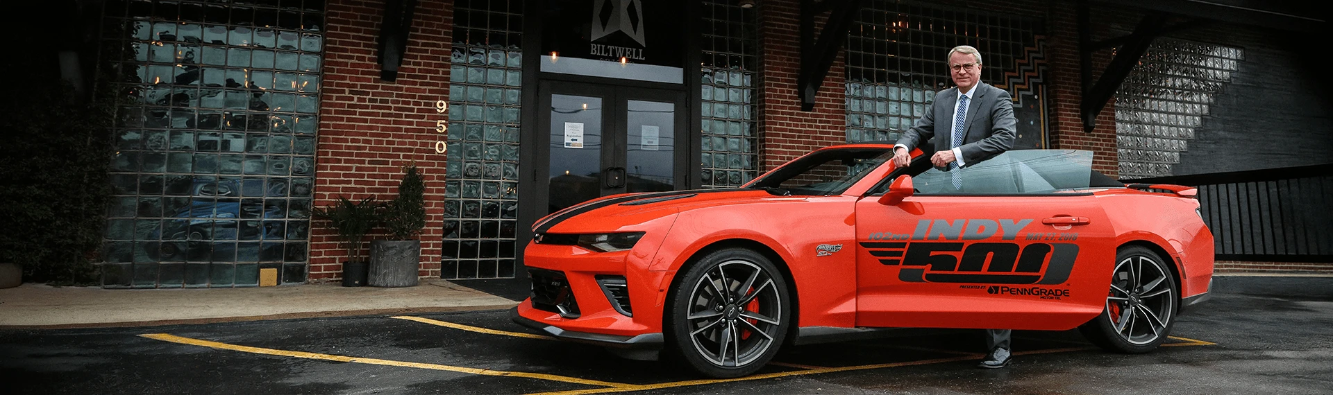 President Elsener in a suit standing beside a bright red convertible Camaro with prominent Indy 500 branding, parked in front of a historic brick building as part of Marian University’s Racing Heritage exhibit.