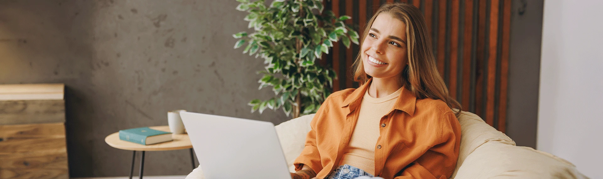 A smiling young woman with long brown hair sits comfortably on a couch with a silver laptop on her lap, wearing an orange shirt and jeans while looking at the camera in a cozy, modern indoor setting with a green plant in the background.