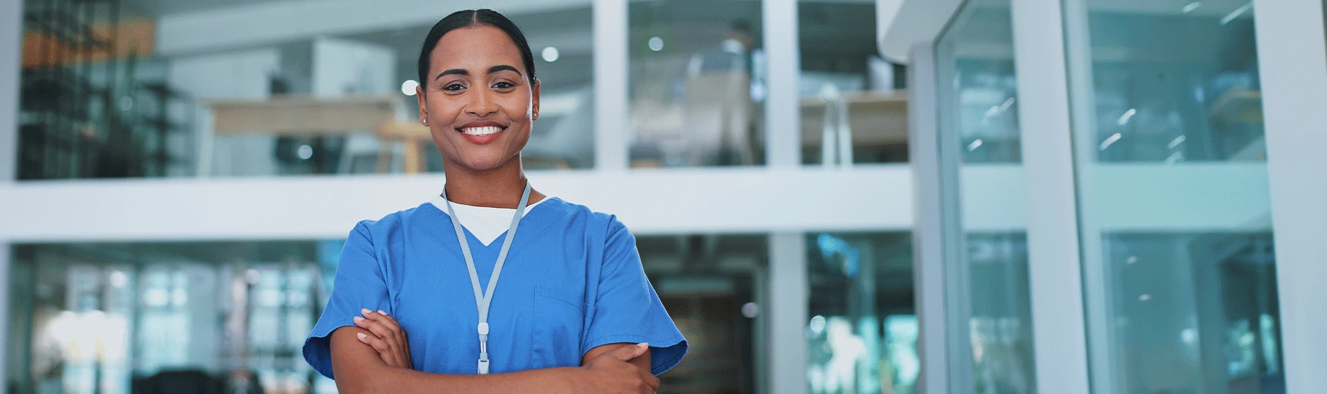A smiling woman in blue scrubs with a lanyard stands confidently with her arms crossed in a modern healthcare setting, looking directly at the camera.