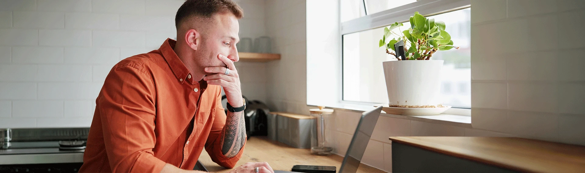 A thoughtful man with short brown hair and a tattoo on his forearm sits at a wooden table in an orange shirt, resting his chin on his hand while looking to the side in a bright indoor setting.