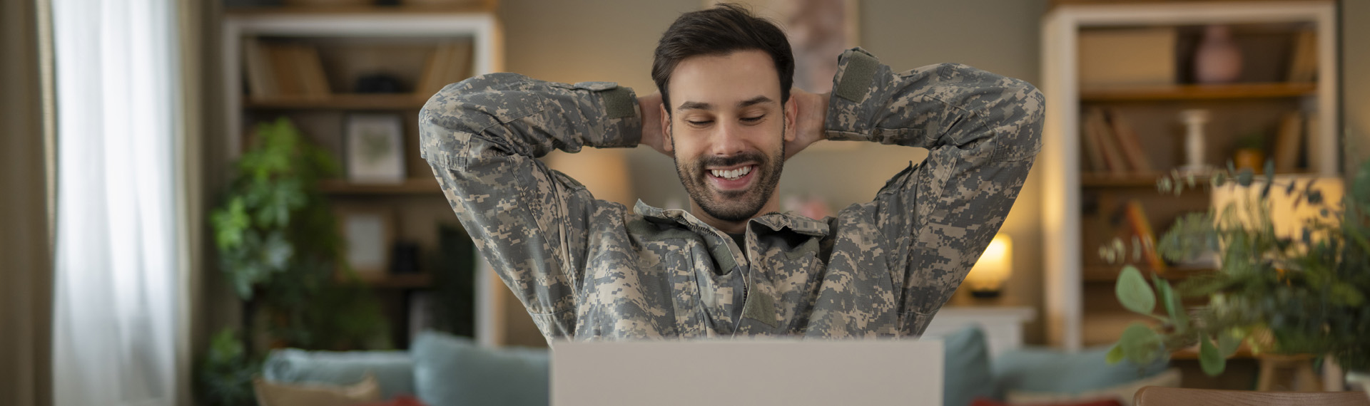 Smiling man in military camouflage uniform relaxing with his hands behind his head while sitting at a laptop at home.