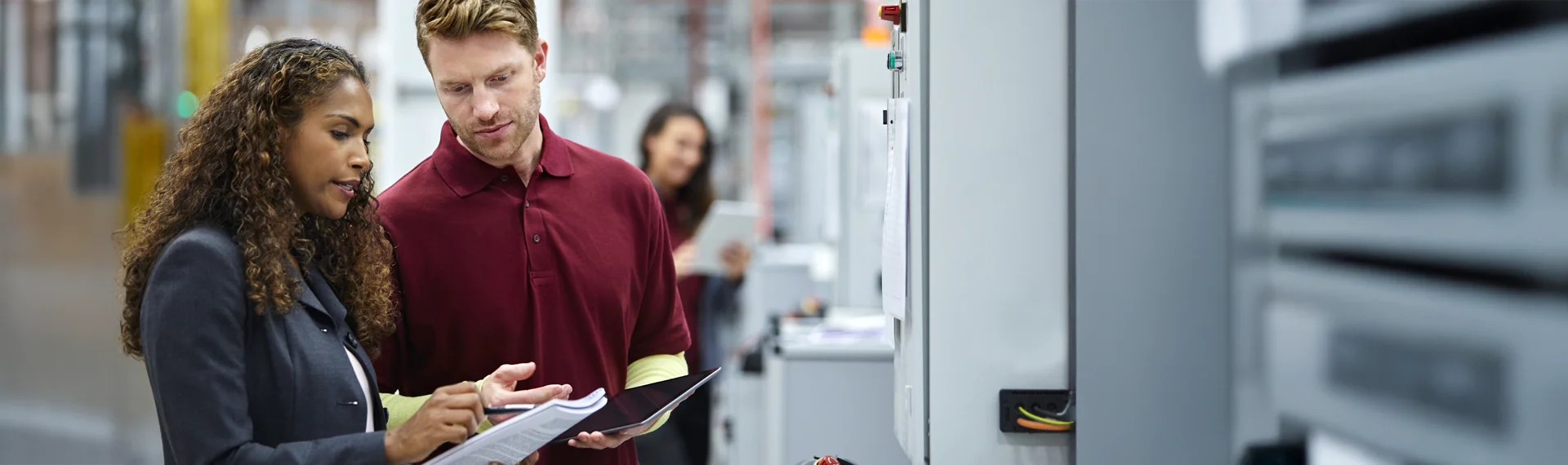 A woman with curly hair wearing a dark blazer and a man in a maroon polo shirt stand side-by-side in a modern industrial or manufacturing facility, intently reviewing documents and a tablet together. The woman points at the materials while the man looks on; another professional is visible working in the background amid control panels and machinery.
