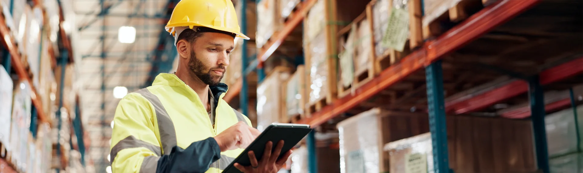 A focused warehouse worker wearing a yellow hard hat and high-visibility safety jacket stands in a large distribution center, using a tablet to manage inventory or logistics operations among tall shelves stocked with goods and pallets.