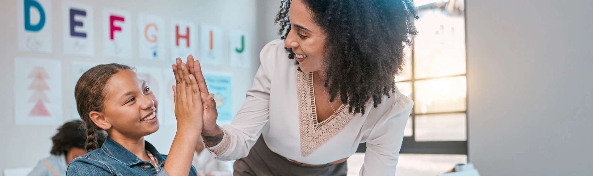 Smiling teacher holding a tablet gives a high five to a young student in a denim jacket at her desk in a classroom with alphabet letters on the wall.