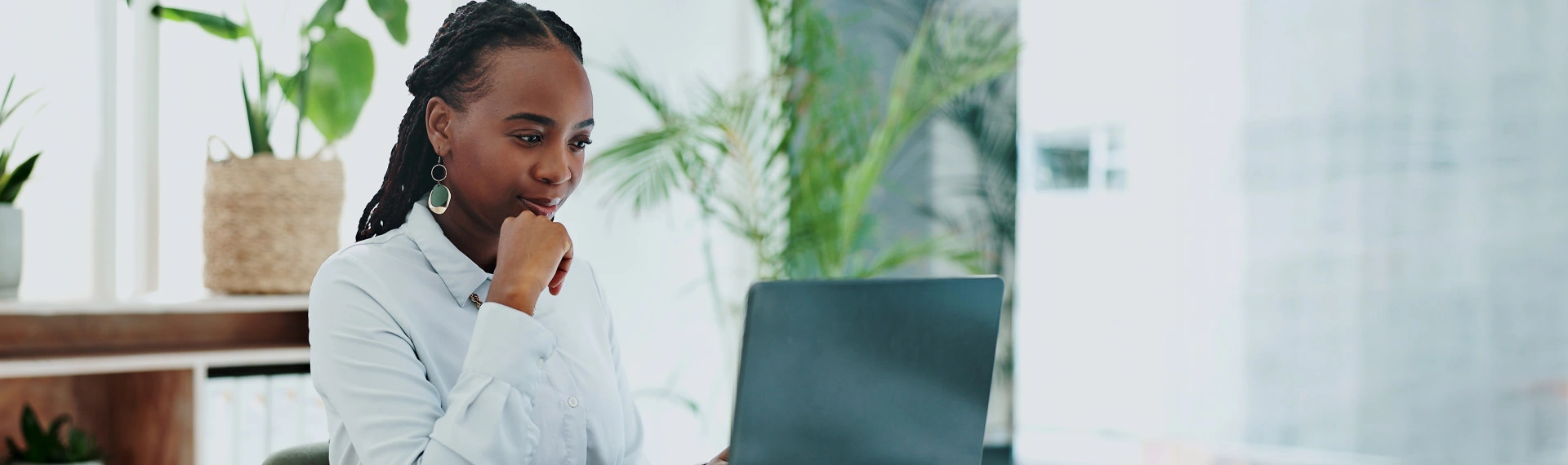 Woman with braided hair and a light button-up shirt rests her chin on her hand while looking thoughtfully at a laptop in a bright office with potted plants.