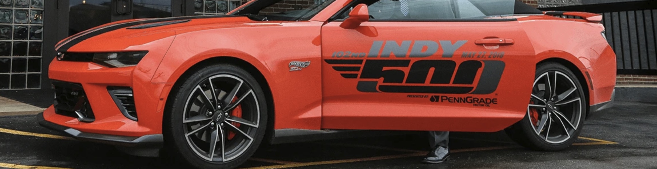 President Elsener in a suit standing beside a bright red convertible Camaro with prominent Indy 500 branding, parked in front of a historic brick building as part of Marian University’s Racing Heritage exhibit.