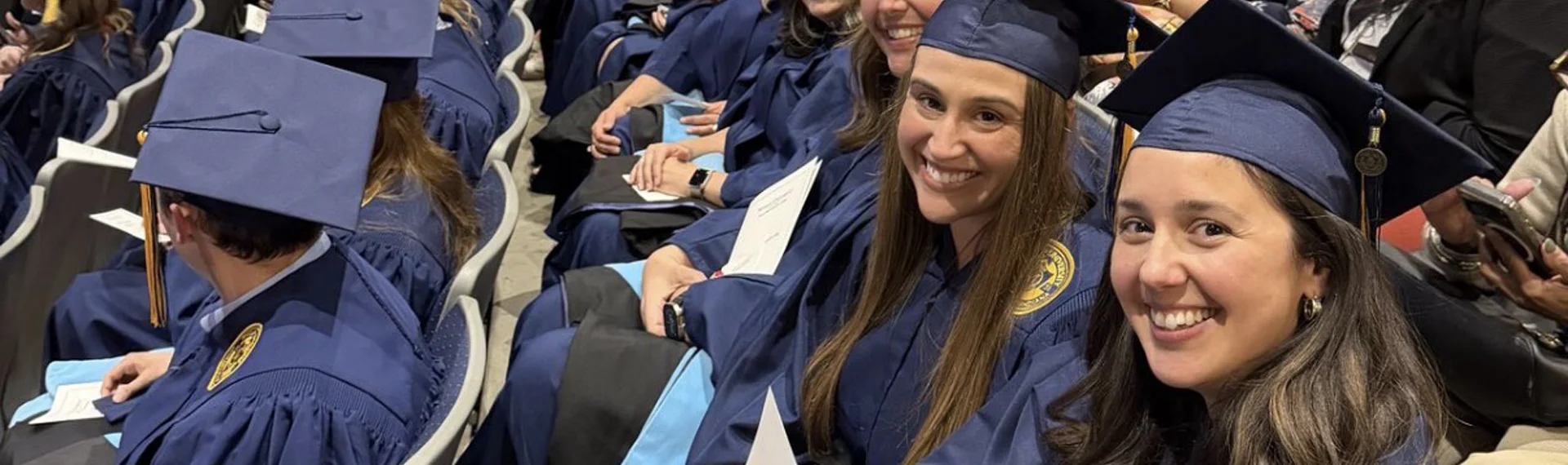 Smiling graduates in navy caps and gowns with light blue hoods sit in rows at a commencement ceremony, holding programs, with two women in the foreground turning toward the camera.