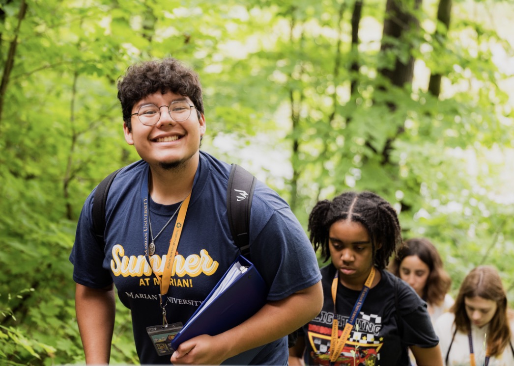 A smiling student hiking on a trail, accompanied by classmates and surrounded by lush greenery.