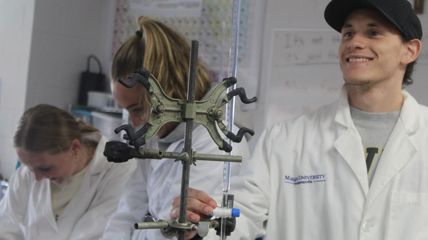 Students in lab coats conducting an experiment, with one student smiling while holding a laboratory apparatus.