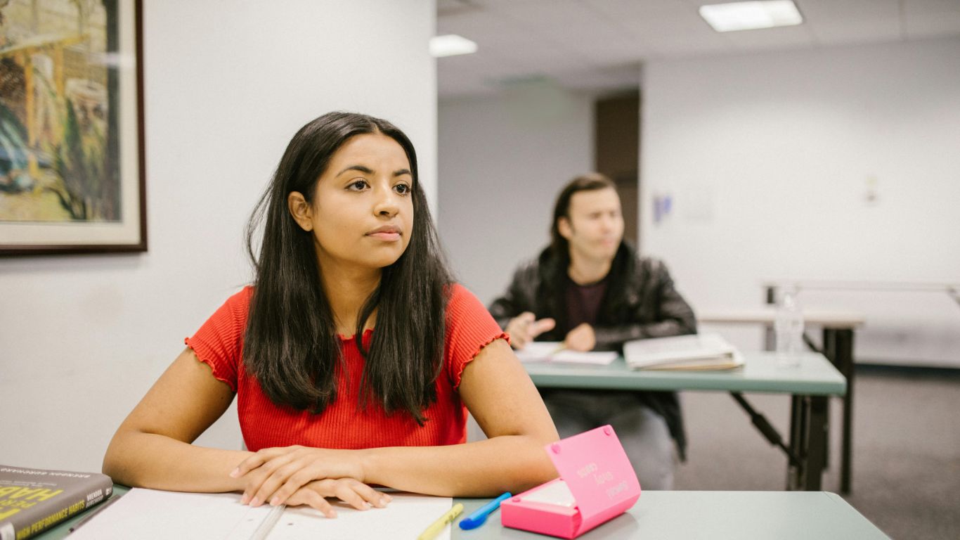 A female student in a red shirt looks thoughtfully while seated at a desk in a classroom, with another student in the background.