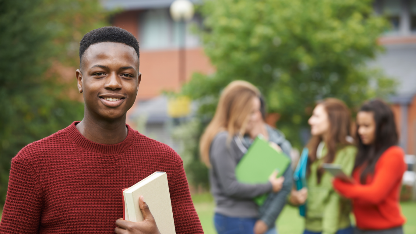 A smiling Black male student holding books, with a group of female students engaged in conversation blurred in the background.