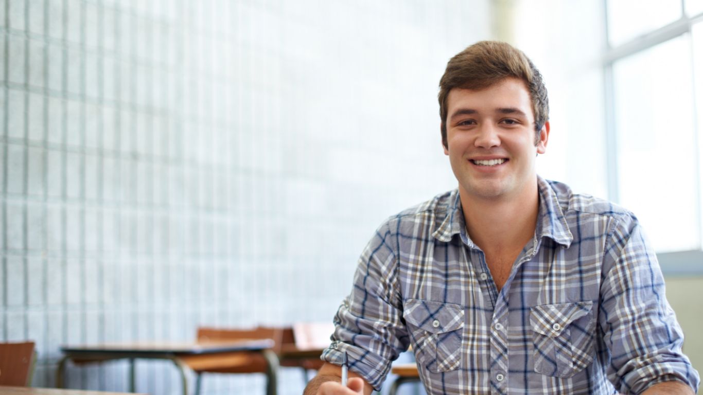 A smiling young man in a plaid shirt sitting at a desk in a classroom setting.