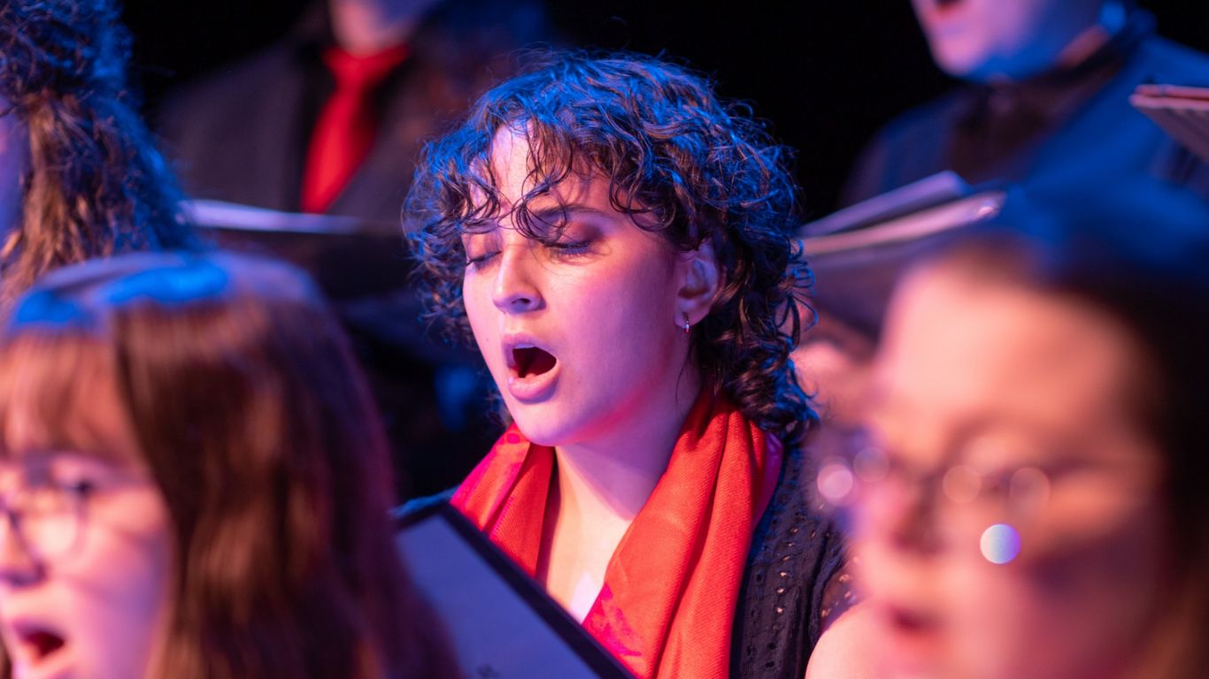 A choir member with curly hair and a red scarf passionately singing while others are blurred in the background.